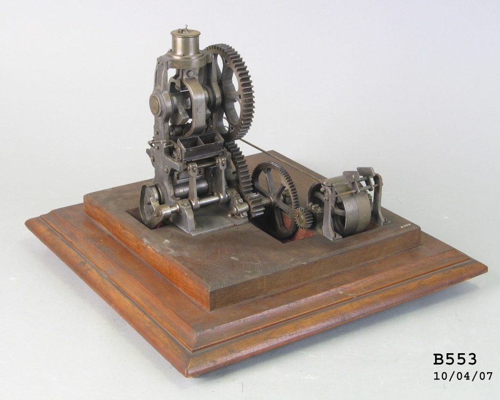 A model of a complex machine with four interlocking gears attached to a set of pistons and two brick shaped moulds. The working model is mounted on an ornate timber base.